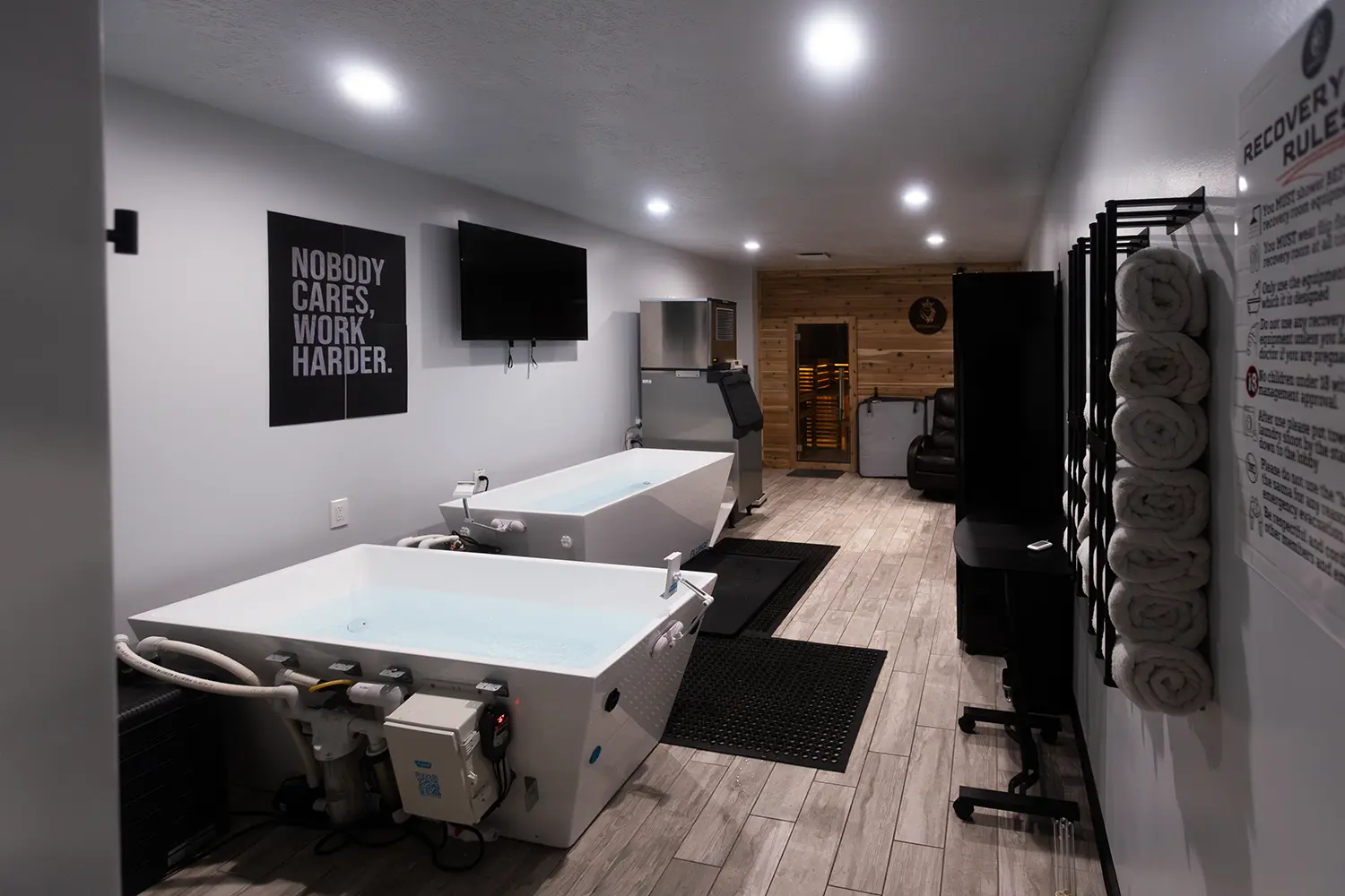 Modern recovery room with two white hydrotherapy tubs filled with water, a sauna at the back, rolled towels on a rack, a black TV mounted on the wall, and motivational sign reading 'Nobody cares, work harder.'
