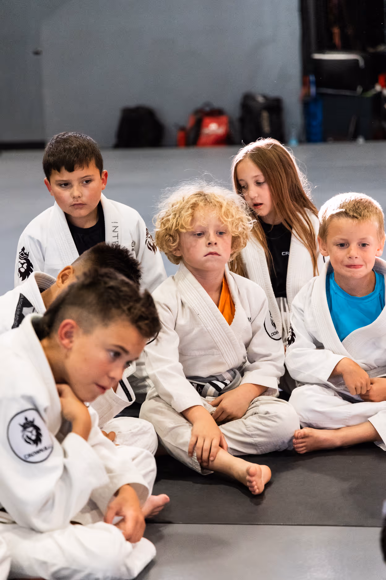 Group of young children in white martial arts uniforms sitting on a mat, listening attentively.