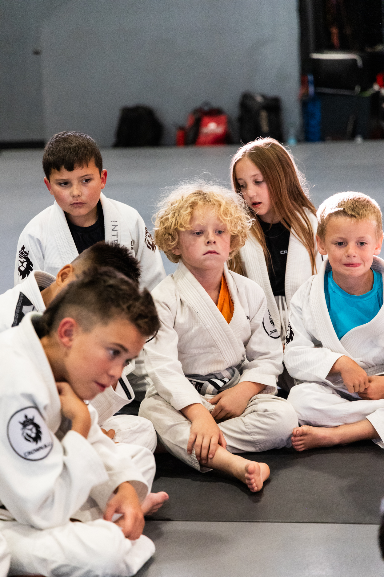 Group of young children in white martial arts uniforms sitting on a mat, listening attentively.