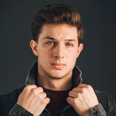 Portrait of a young man with short brown hair holding the collar of his dark jacket against a dark background.