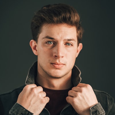 Portrait of a young man with short brown hair holding the collar of his dark jacket against a dark background.