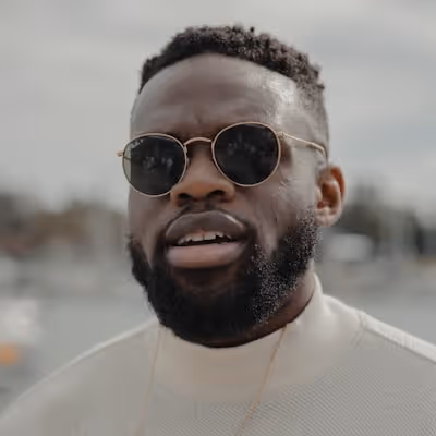 Close-up of a man with a beard wearing round sunglasses and a light sweater outdoors.