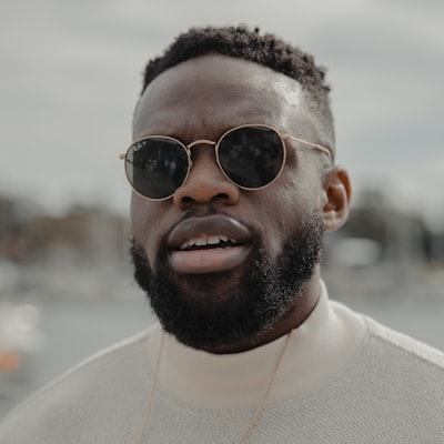 Close-up of a man with a beard wearing round sunglasses and a light sweater outdoors.