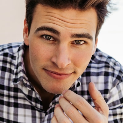 Close-up of a young man with dark hair wearing a black and white checkered shirt, looking thoughtfully at the camera and touching his chin.