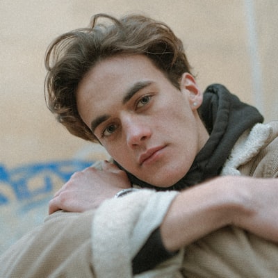 Young man with wavy hair resting his head on his crossed arms, wearing a beige jacket over a black hoodie.