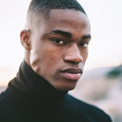 Portrait of a young man with short hair wearing a black turtleneck, looking thoughtfully to the side against a blurred outdoor background.
