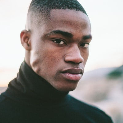 Portrait of a young man with short hair wearing a black turtleneck, looking thoughtfully to the side against a blurred outdoor background.