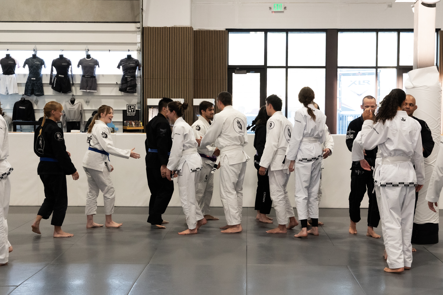 Group of barefoot adults in white and black martial arts uniforms greeting each other inside a dojo with martial arts apparel displayed on the wall.