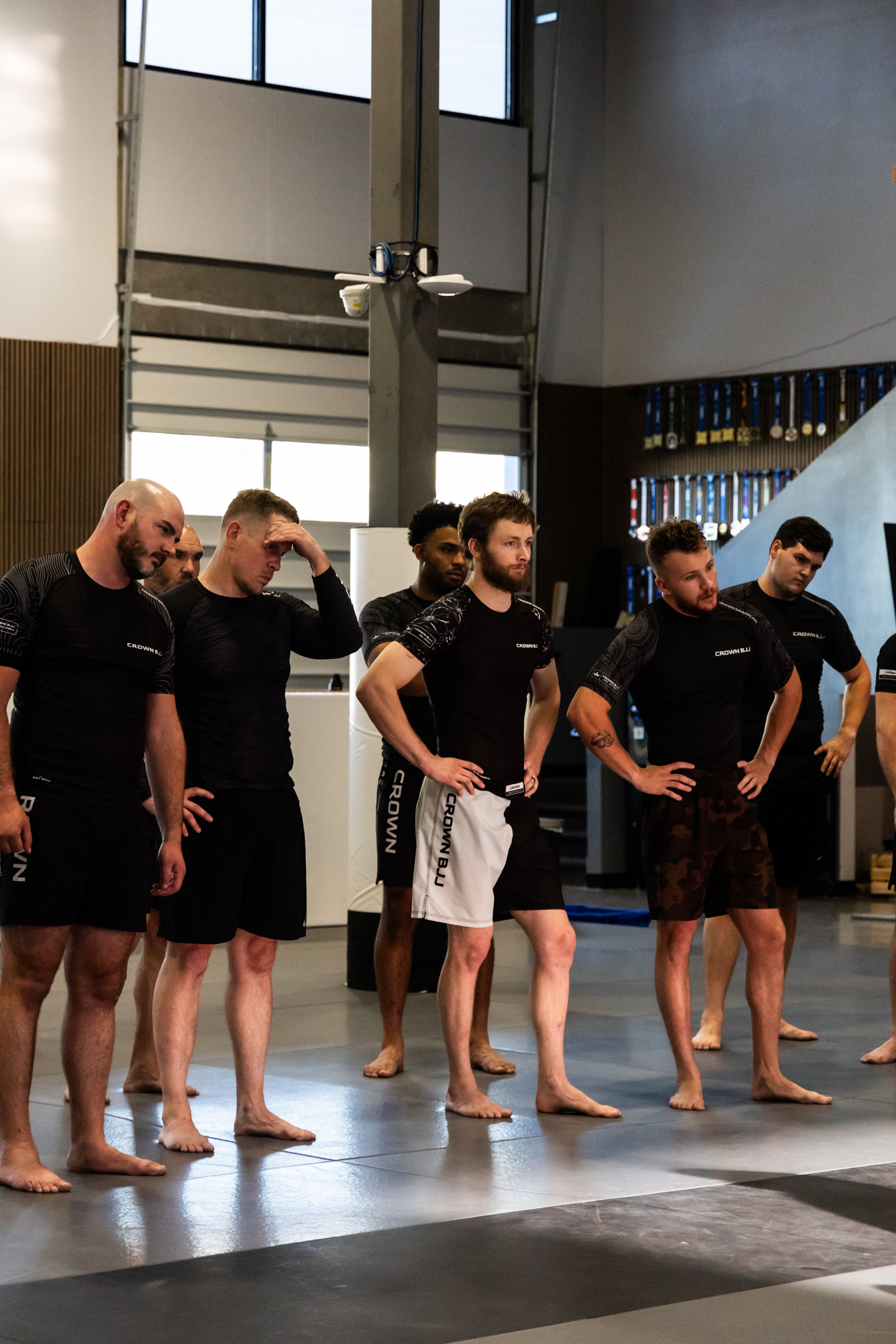 Group of barefoot men in black Crown BJJ training attire standing on mats in a martial arts gym.
