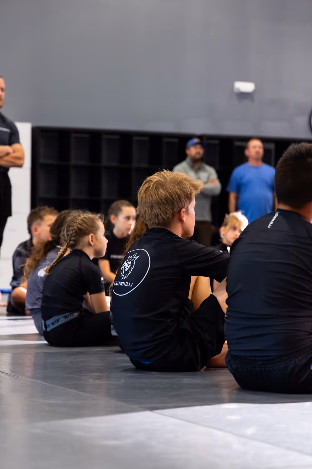 Children sitting on a mat in a gym listening attentively, some wearing black shirts with a Crown BJJ logo.