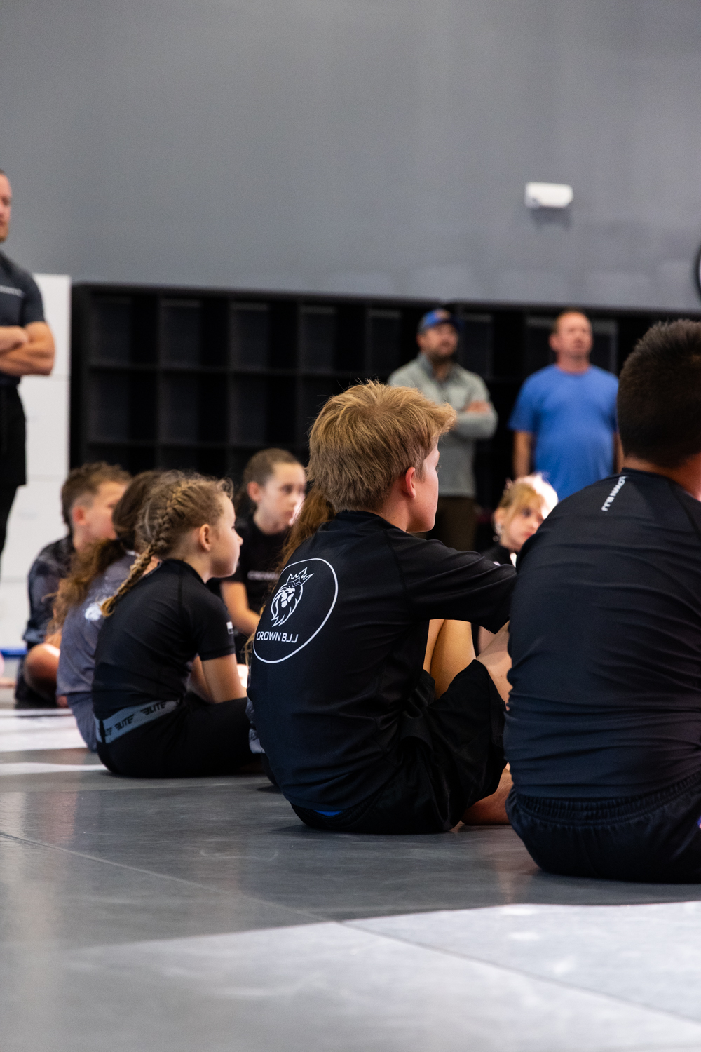 Children sitting on a mat in a gym listening attentively, some wearing black shirts with a Crown BJJ logo.