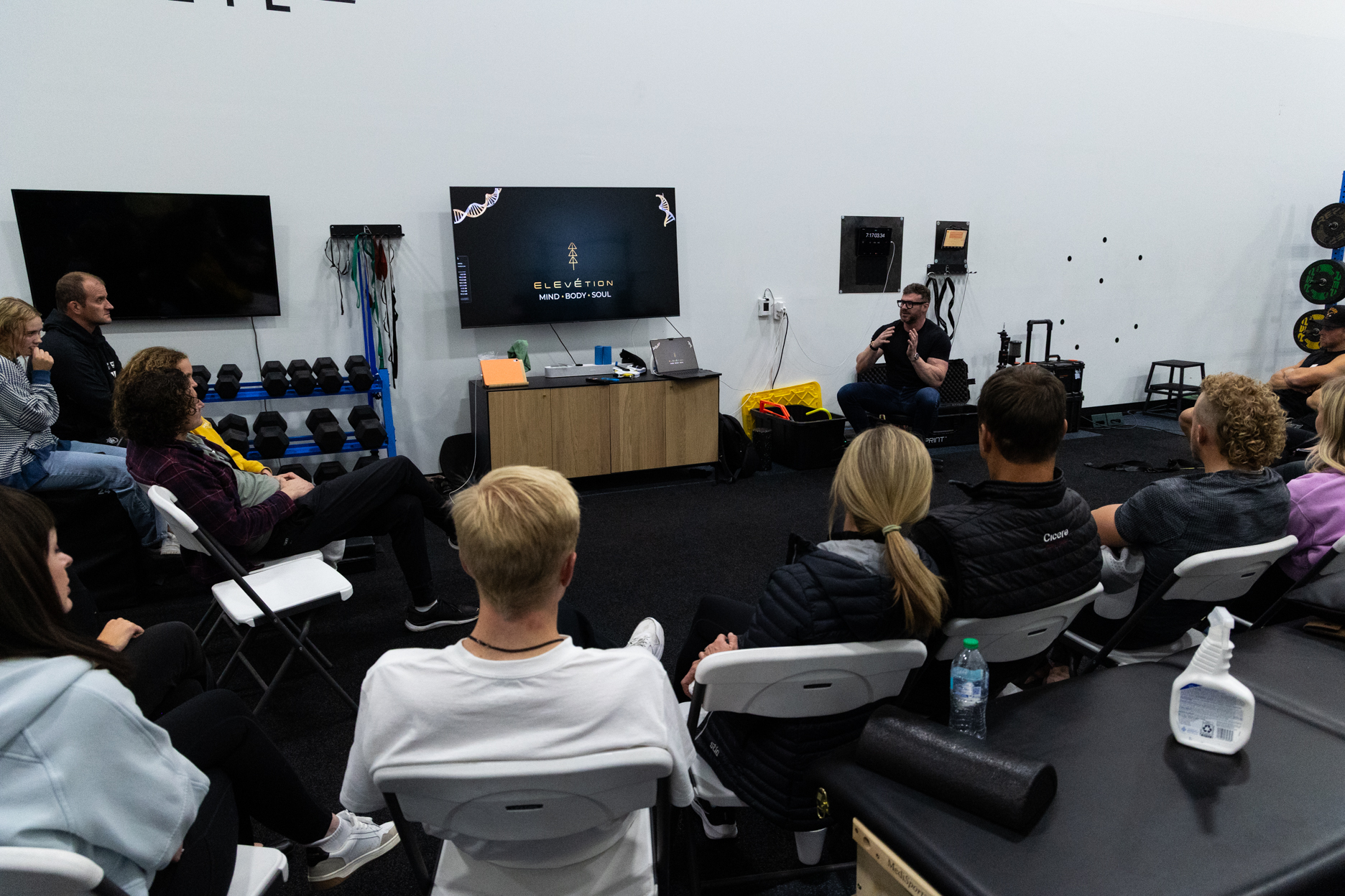 Group of people seated in folding chairs attentively listening to a speaker in a gym setting with exercise equipment around.