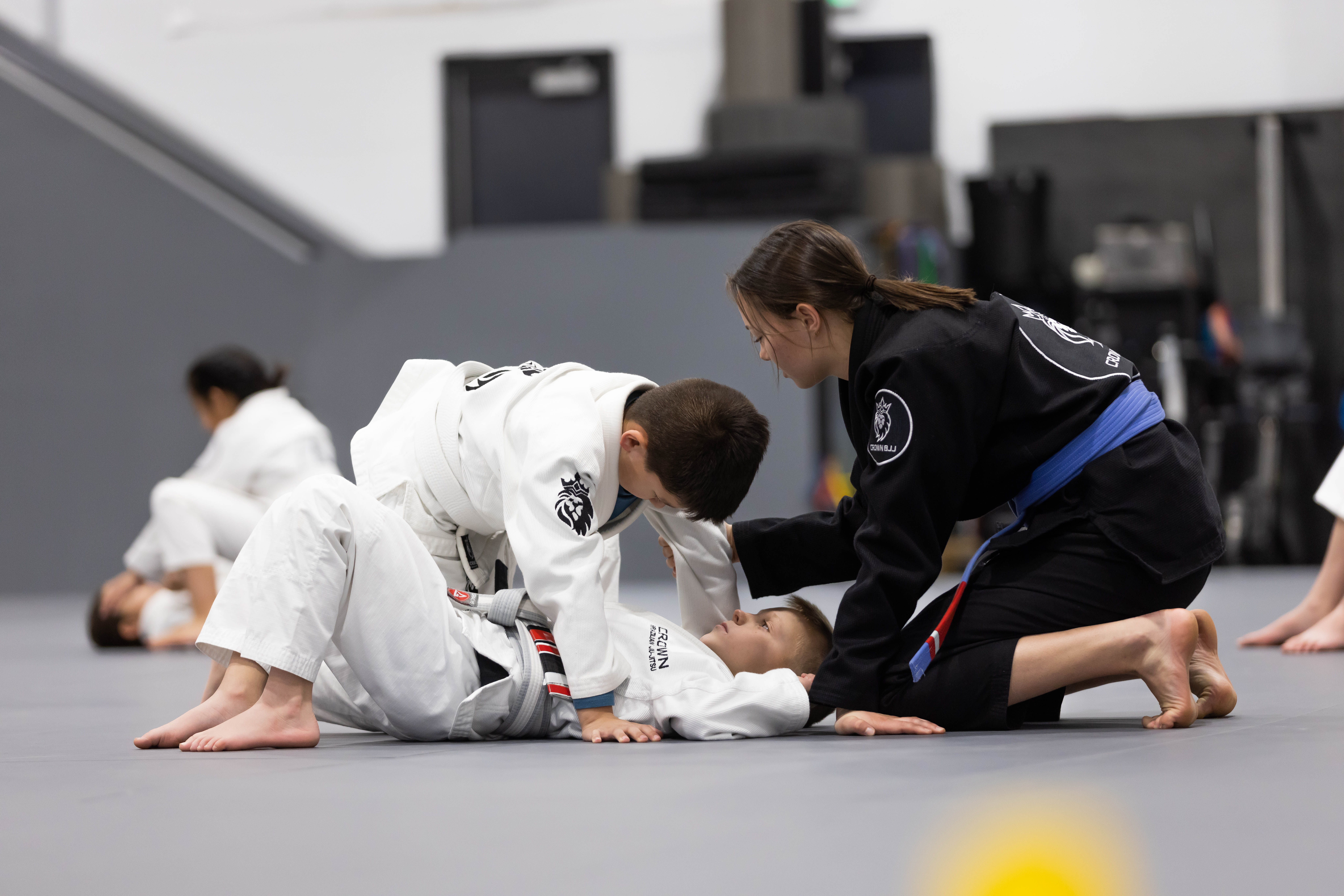 A female instructor in a black gi with a blue belt teaches Brazilian Jiu-Jitsu to two boys in white gis practicing a grappling technique on the mat.