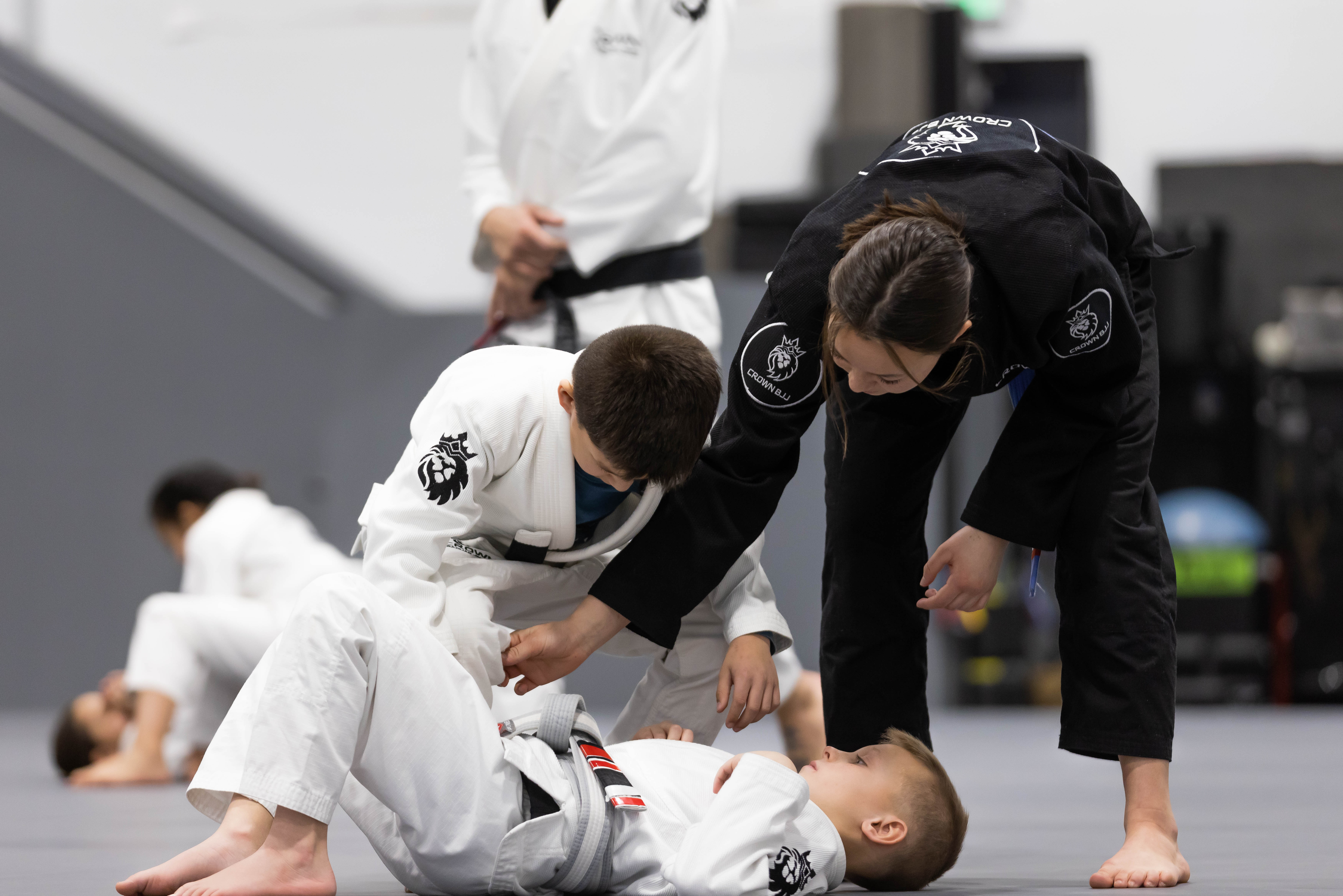 Children practicing Brazilian Jiu-Jitsu on mats, with one instructor guiding a young boy in a white gi while another child lies on the ground.
