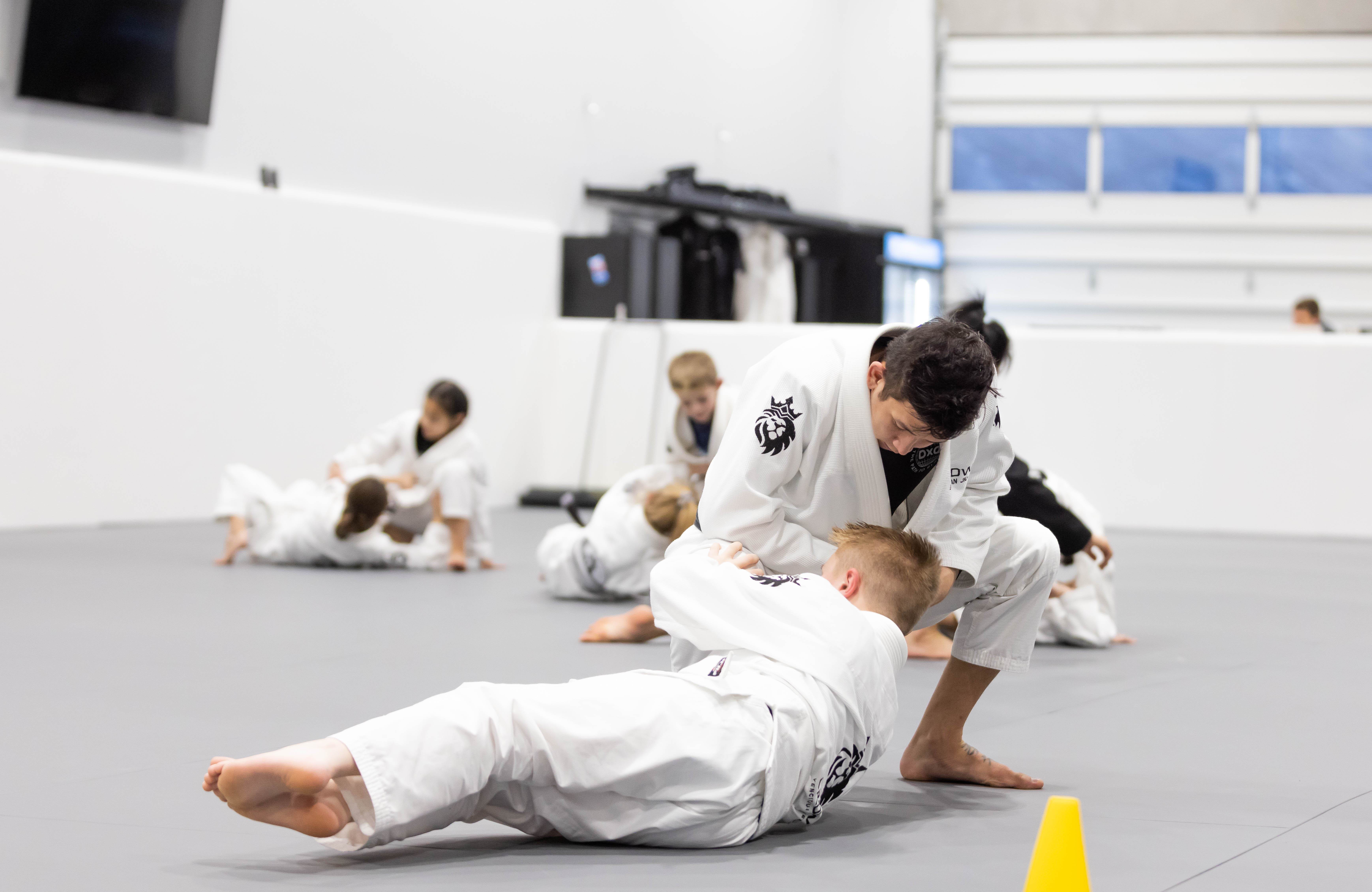 Two children practicing Brazilian Jiu-Jitsu on mats in a training gym while others train in the background.