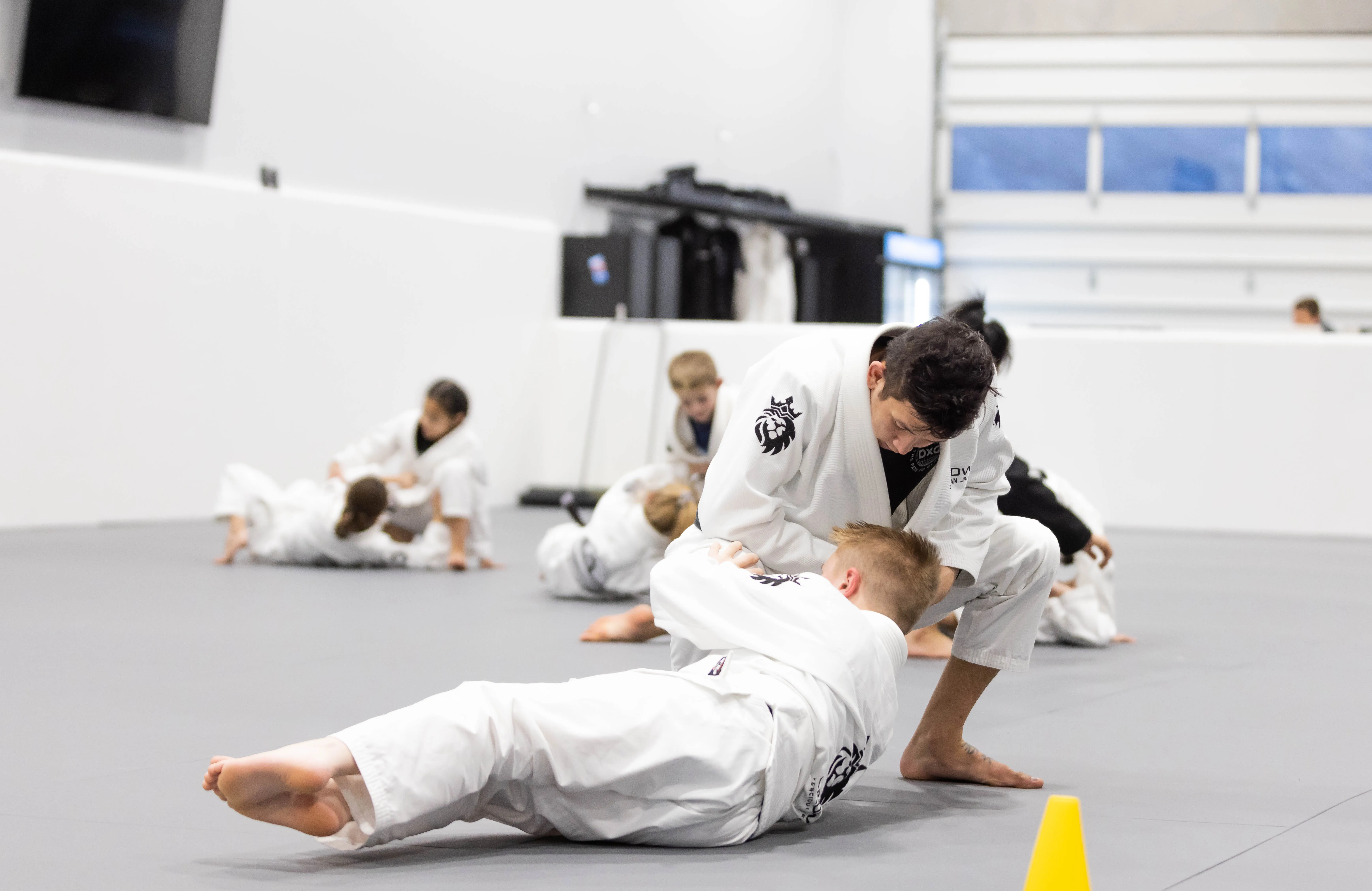 Two children practicing Brazilian Jiu-Jitsu on mats in a training gym while others train in the background.