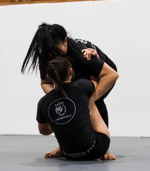 Two people practicing Brazilian Jiu-Jitsu wearing black rash guards, one sitting with legs wrapped around the other's torso in a gym setting.