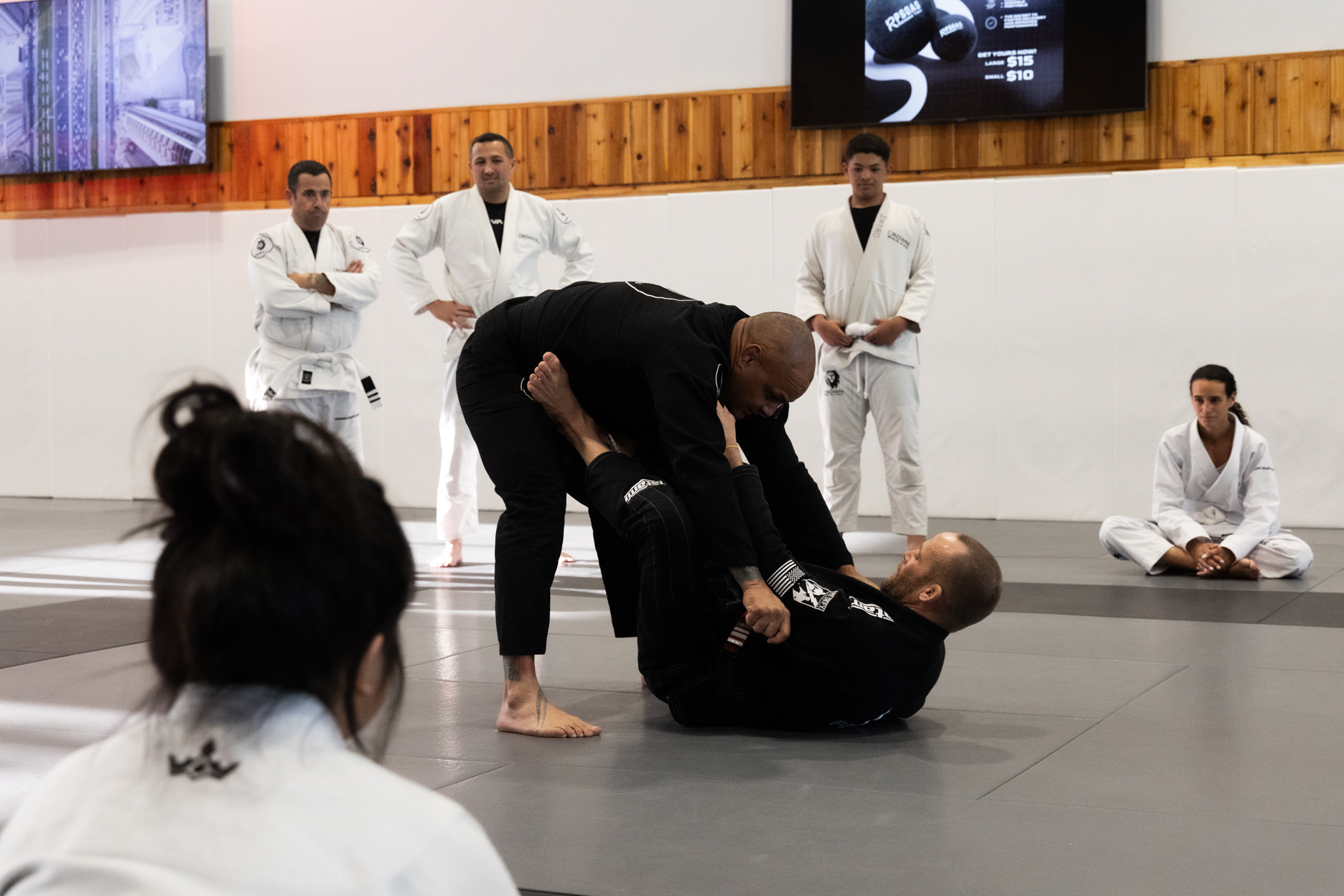 Two men practicing Brazilian Jiu-Jitsu on mats while others in white gis observe.