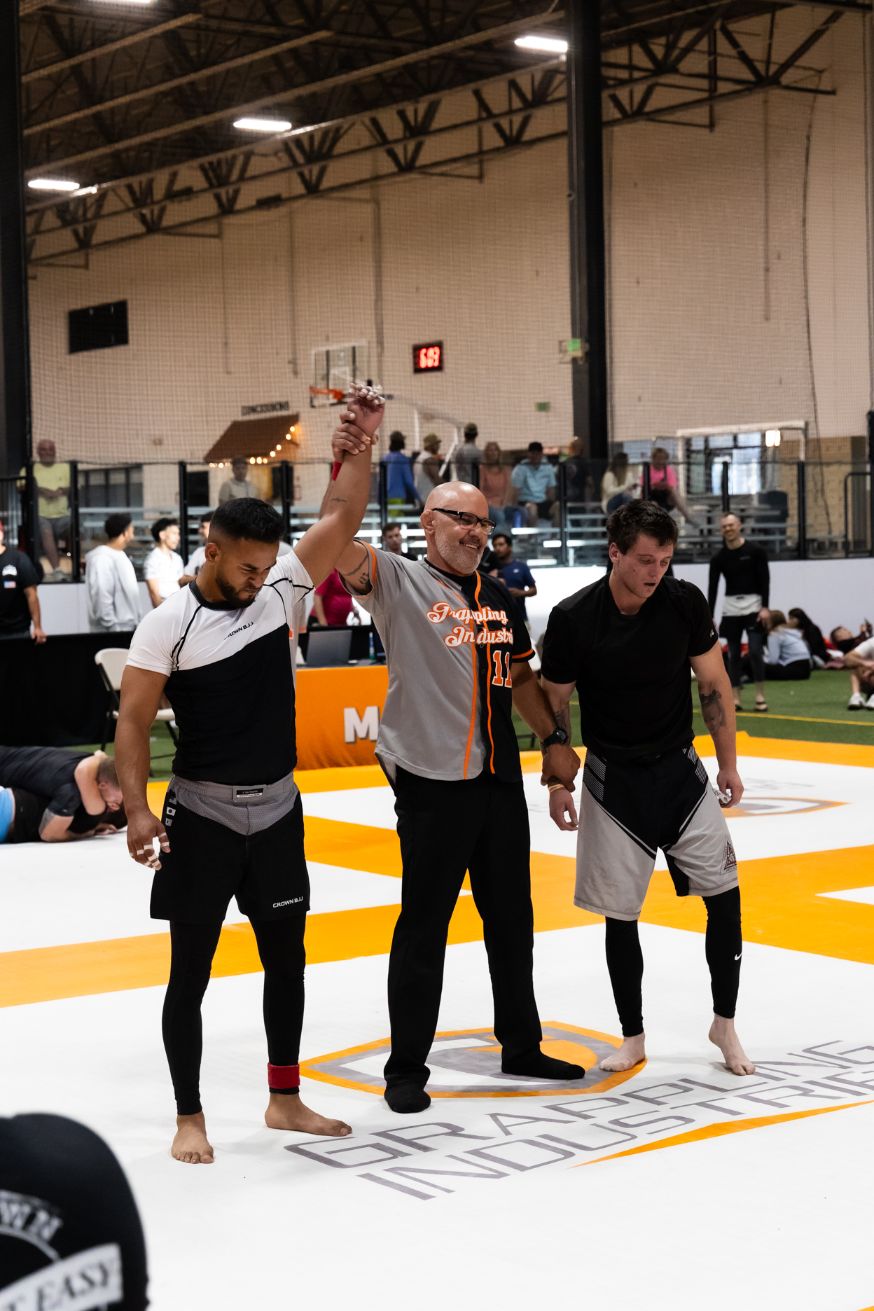 Referee raising the arm of a victorious male grappler in a sports arena with spectators in the background.