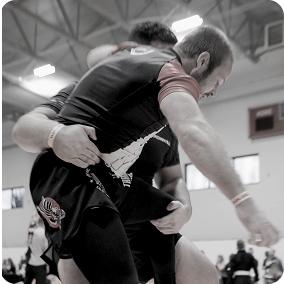 Two men engaged in a grappling martial arts match inside a gymnasium.