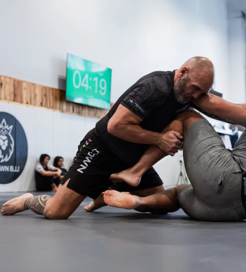 Two men engaged in Brazilian Jiu-Jitsu grappling on a mat in a gym with a timer displaying 04:19 in the background.