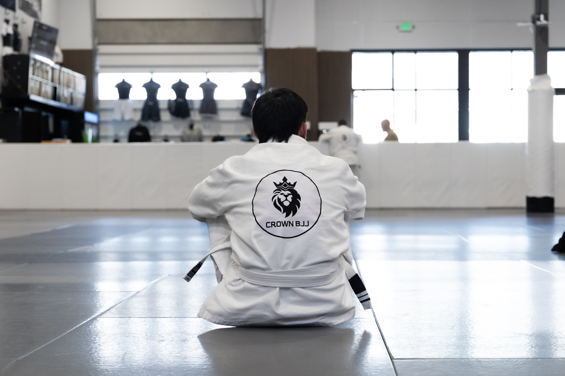 Person in a white Brazilian Jiu-Jitsu gi sitting on a mat facing away, with Crown BJJ logo on the back.