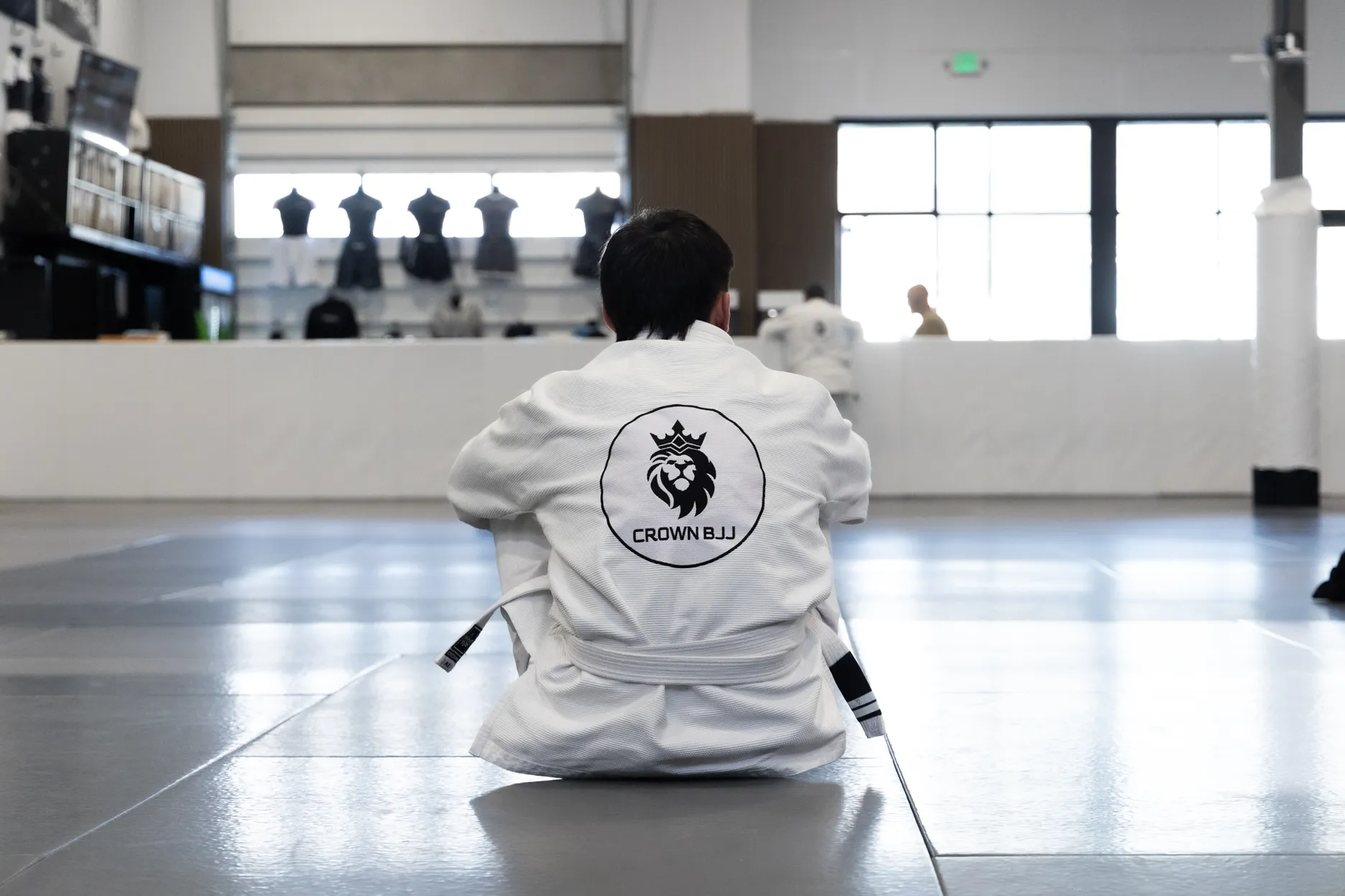 Person in a white Brazilian Jiu-Jitsu gi sitting on a mat facing away, with Crown BJJ logo on the back.