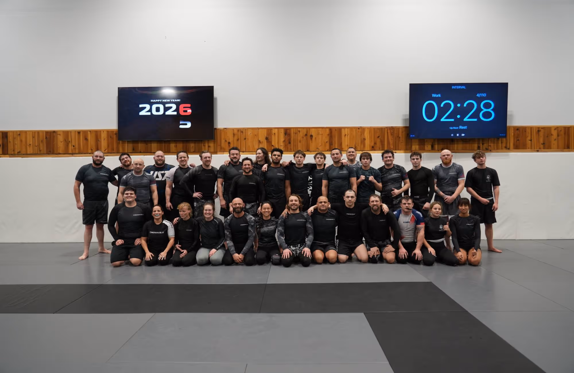 Group of martial artists in black training gear posing for a photo in a gym with two screens displaying 'Happy New Year 2025' and a workout timer.