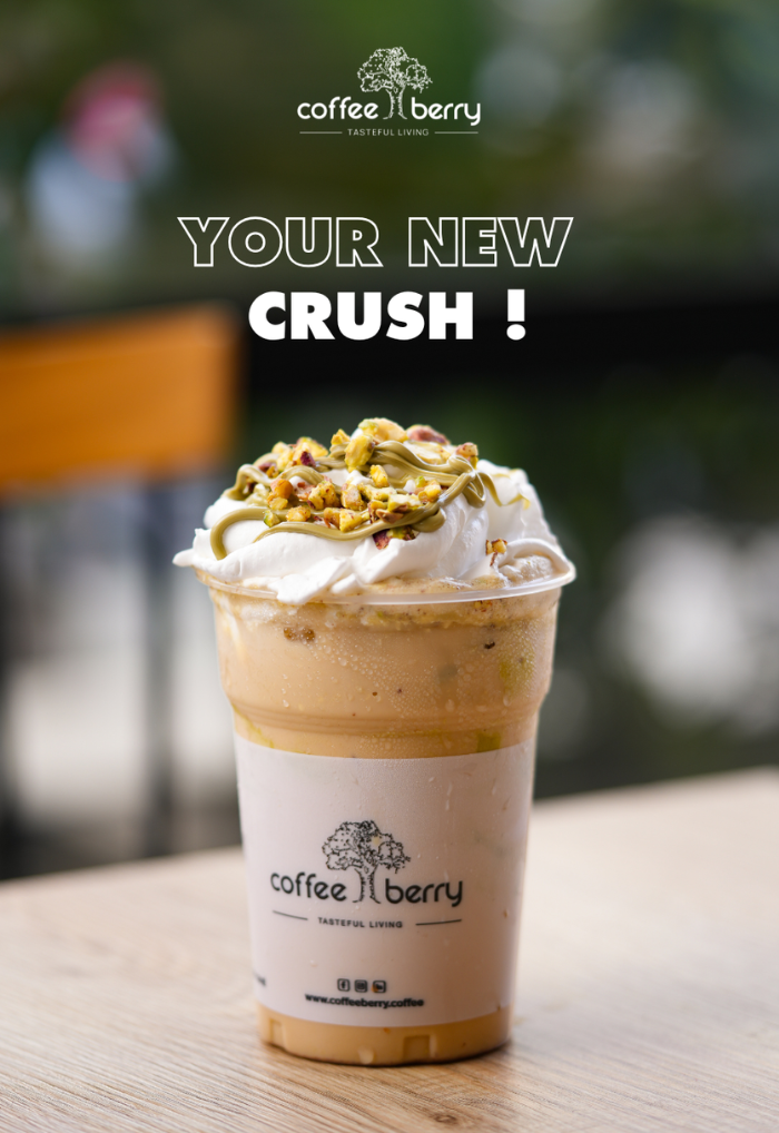 Iced coffee drink topped with whipped cream, pistachio cream, and crushed nuts in a Coffee Berry branded cup on a wooden table.
