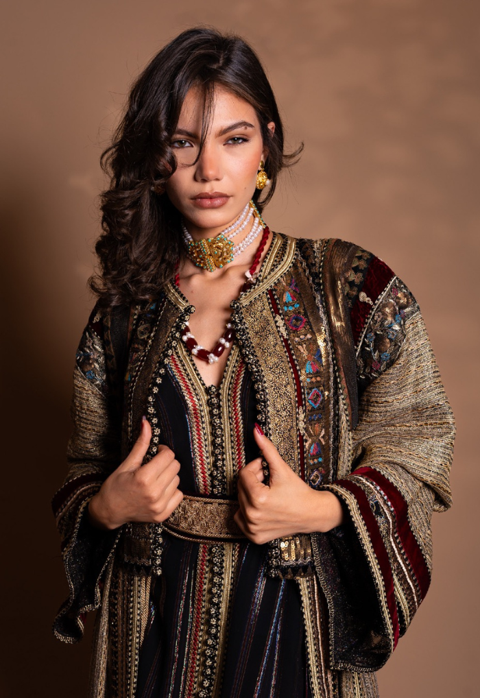 Woman wearing an ornate traditional dress with gold and red embroidery and pearl necklace, posing against a neutral brown background.