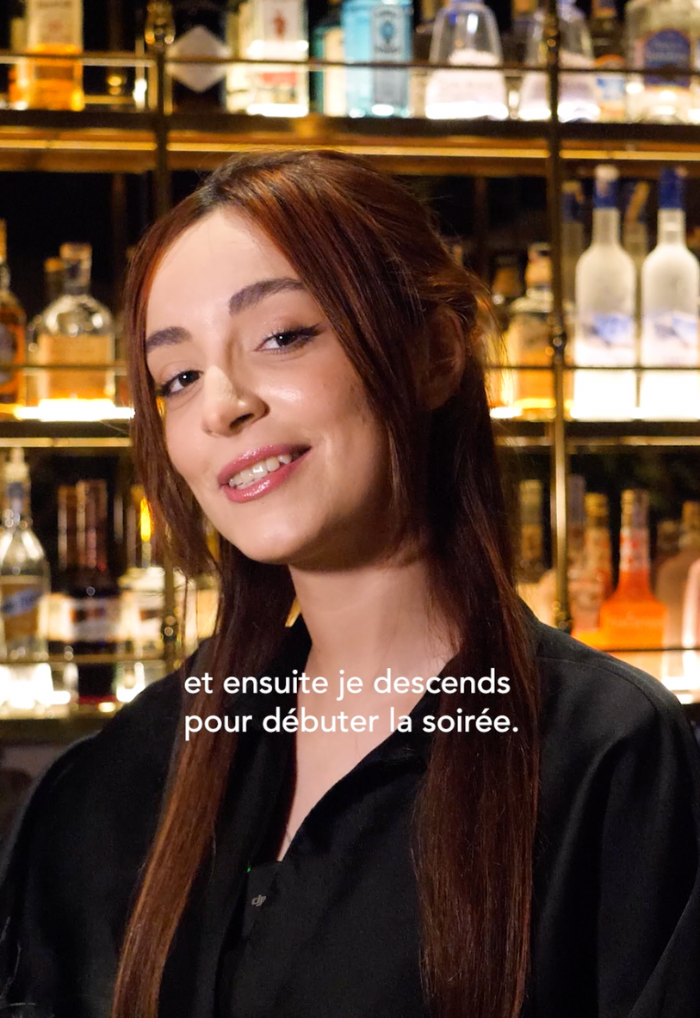 Smiling woman with long brown hair standing in front of a bar shelf with bottles, French subtitle reads 'et ensuite je descends pour débuter la soirée.'