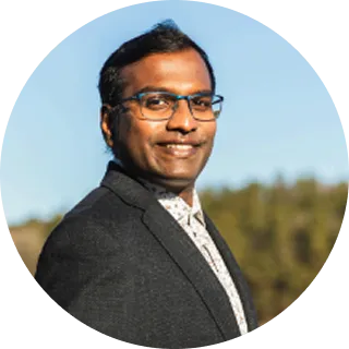 Smiling man wearing glasses and a dark blazer outdoors with trees and blue sky in the background.