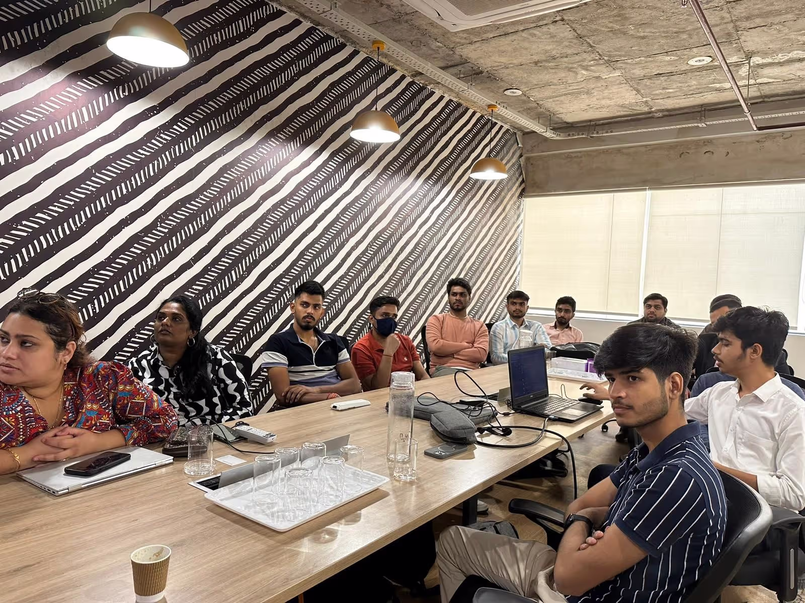 Group of people seated around a table in a modern conference room with a black and white diagonal striped wall and ceiling lights.