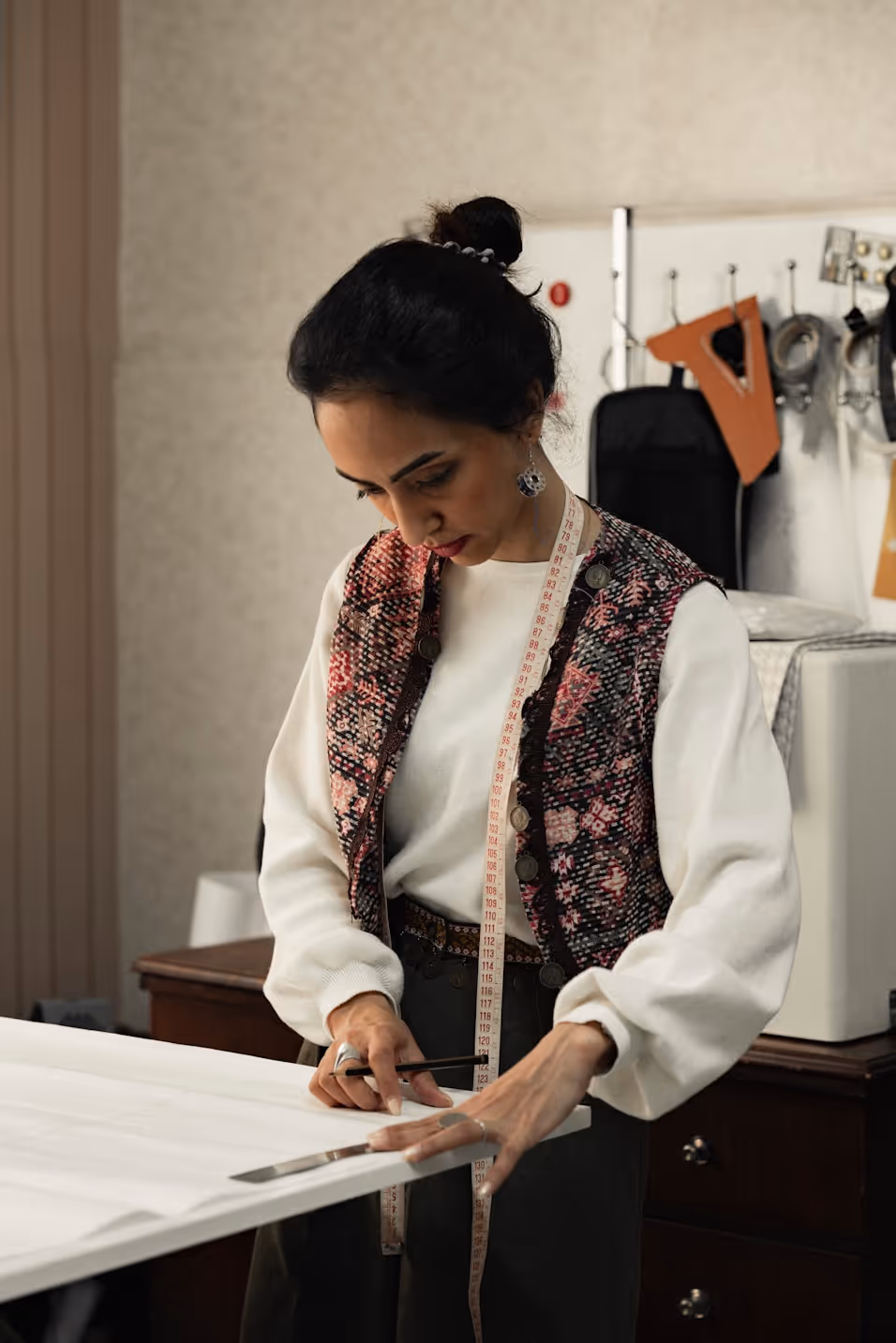 Woman with a measuring tape around her neck marking fabric with a pencil in a sewing workspace.