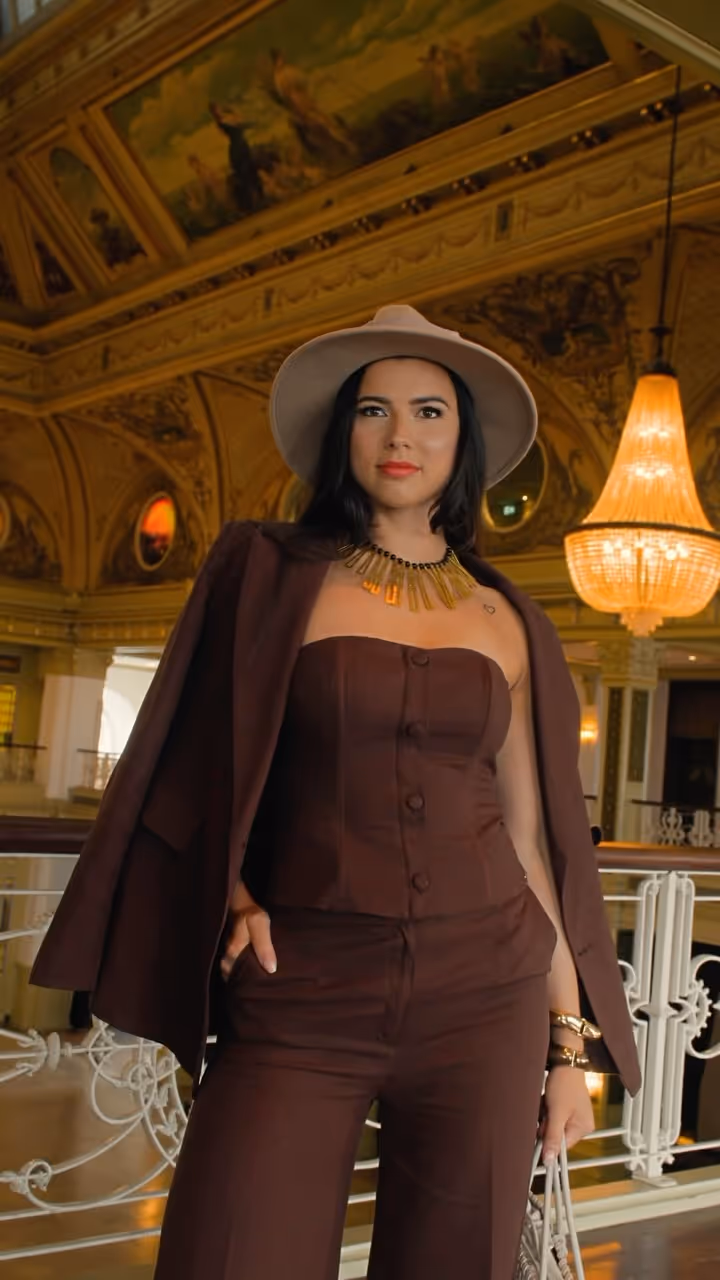 Woman wearing a brown strapless top and matching pants with a brown jacket draped over her shoulders and a beige wide-brimmed hat in an ornate interior with chandeliers.