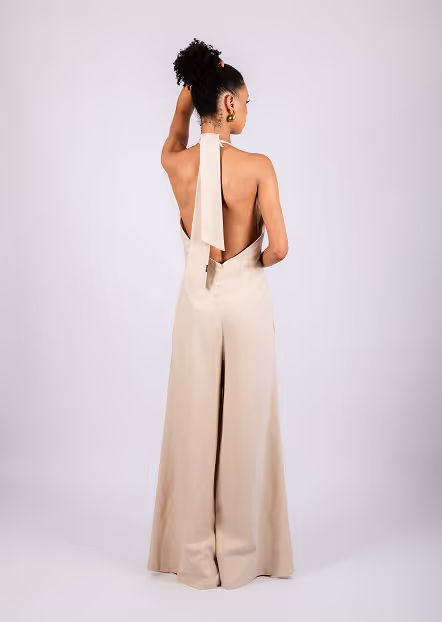Woman with curly hair tied up wearing a backless beige halter jumpsuit with wide legs, standing with her back to the camera.