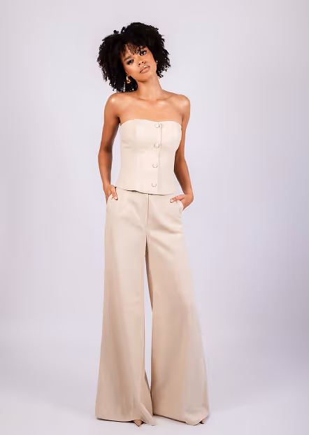 Woman with curly hair wearing a beige strapless top and wide-leg pants, posing with hands in pockets.