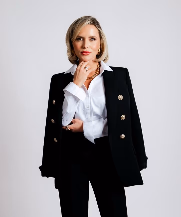 Confident woman with blonde hair wearing a black blazer with gold buttons over a white shirt, accessorized with gold jewelry, standing against a light grey background.