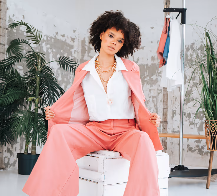 Woman wearing a pink suit and white blouse seated on a white wooden box in a room with potted plants and hanging clothes.