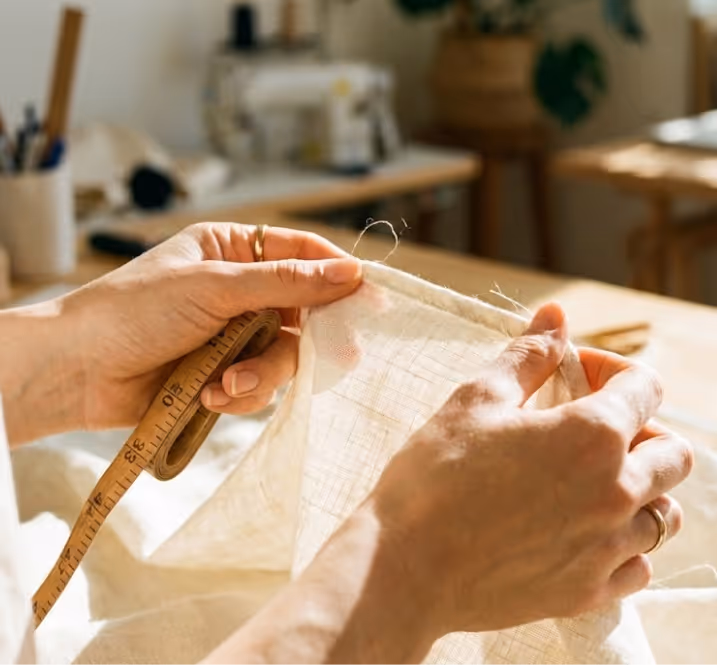 Hands holding and measuring a piece of beige linen fabric with a measuring tape in a sewing room.