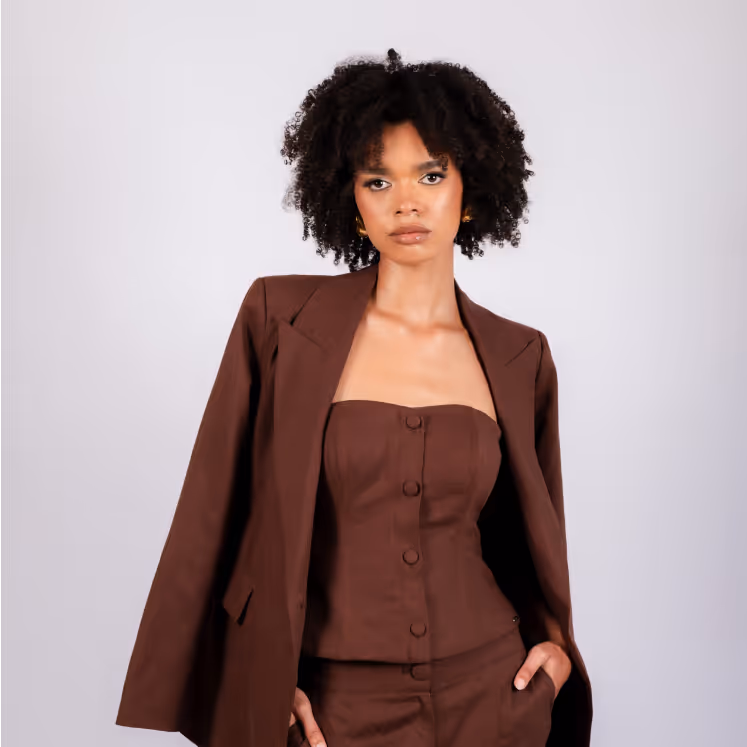 Woman with curly hair wearing a brown strapless top and matching blazer draped over her shoulders against a light background.