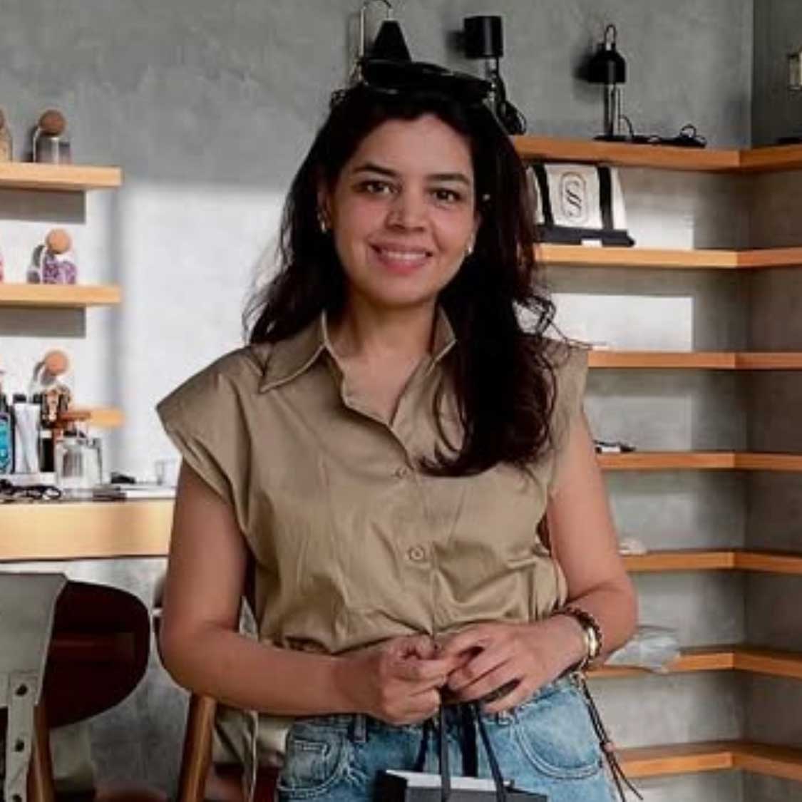 Smiling woman with long dark hair wearing a sleeveless beige shirt and jeans, standing indoors with wooden shelves in the background.