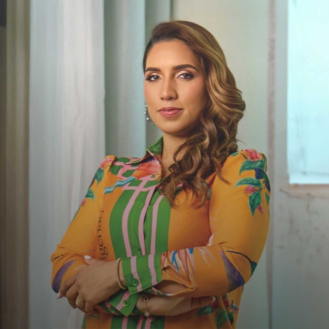 Confident woman with long curled hair wearing a colorful patterned blouse with arms crossed.