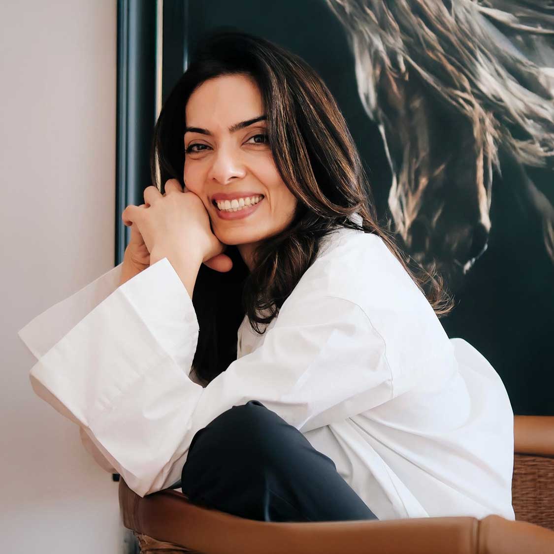 Smiling woman with dark hair wearing a white shirt and sitting with hands clasped near her face.