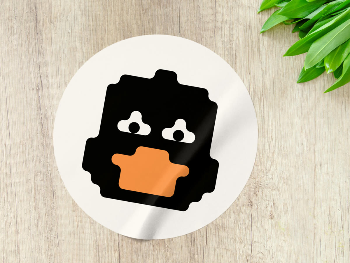 Round sticker on wooden surface featuring a stylized black duck face with white eyes and an orange beak, with green leaves in the top right corner.
