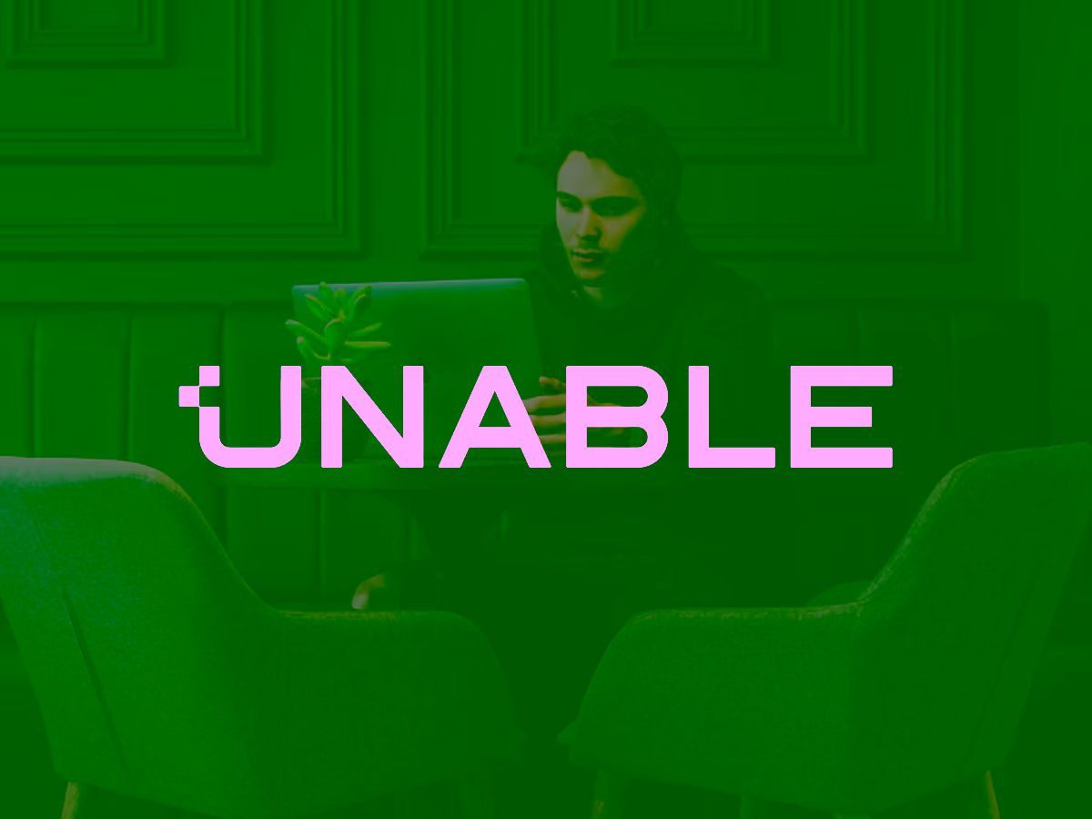 Young man focused on a laptop screen sitting at a table between two gray chairs in a dark blue room with panelled walls.