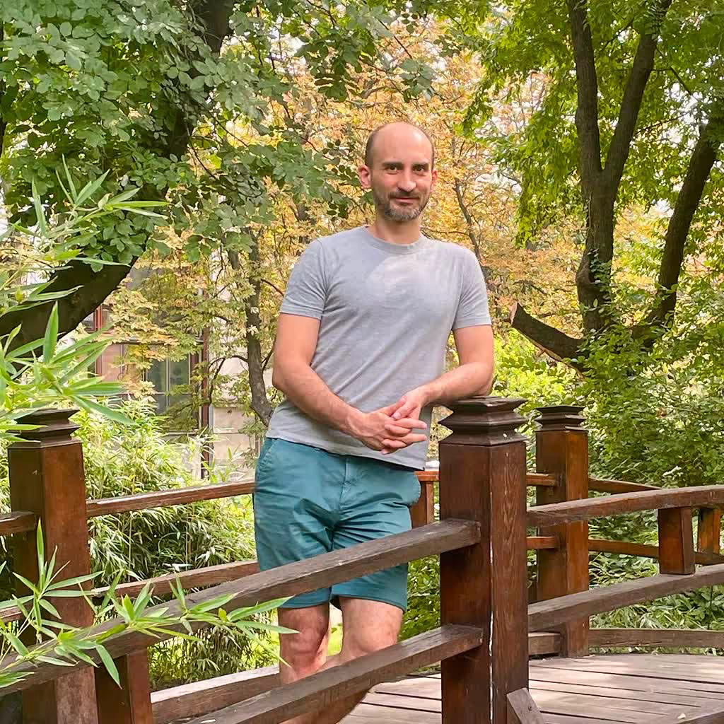 Zlatko Najdenovski, in a grey t-shirt and green shorts, leaning on a wooden railing surrounded by green trees and foliage.