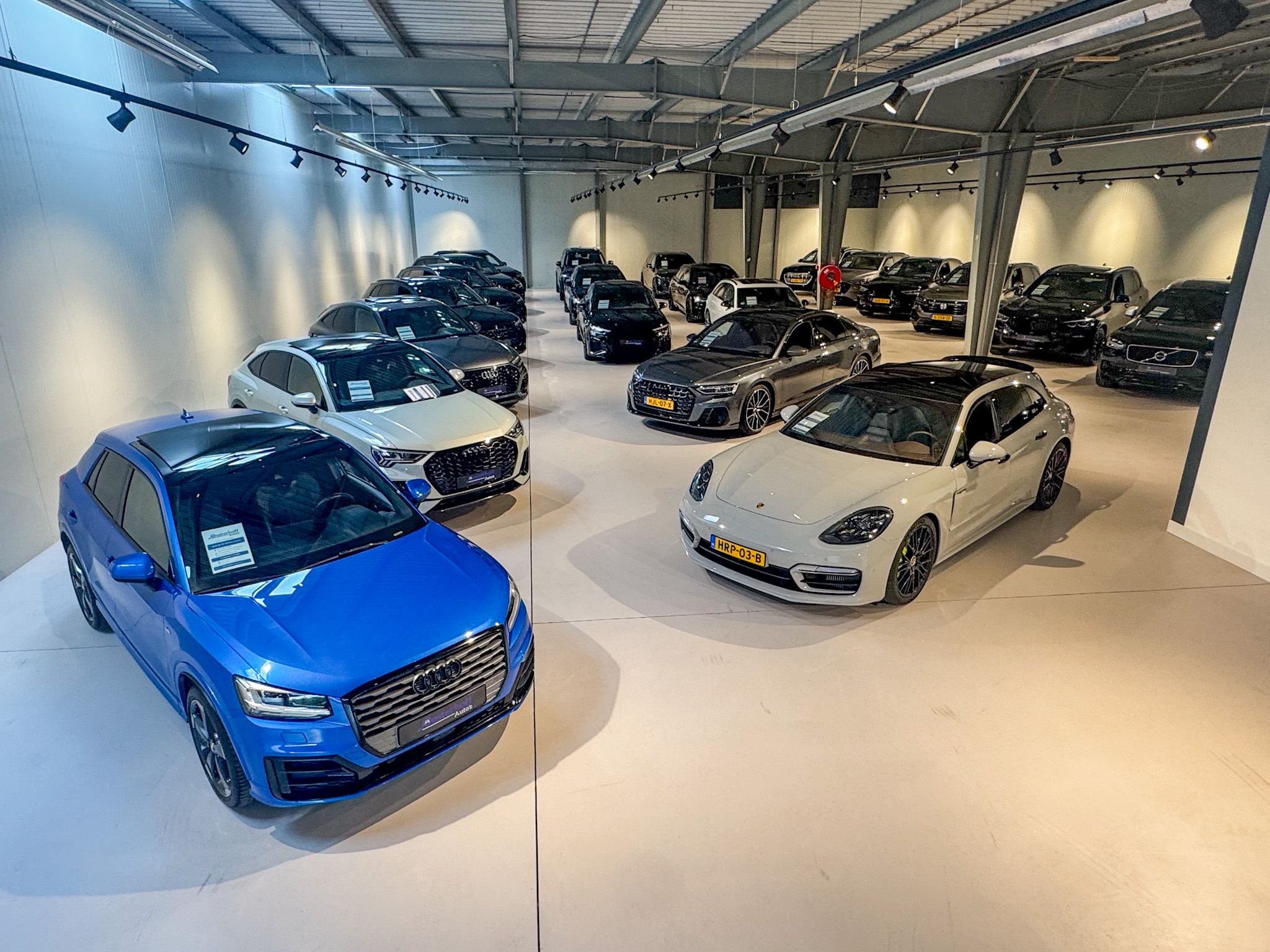 Indoor showroom with multiple parked luxury cars including blue and white Audis and a white Porsche.