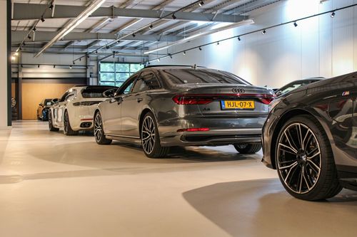 Row of luxury cars parked indoors, featuring a grey Audi A8 with Dutch license plate and a black BMW M series wheel in the foreground.