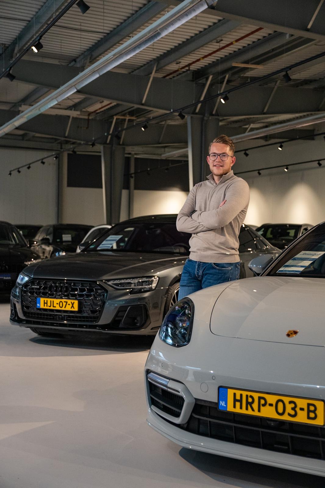 Man with glasses and beige sweater standing with arms crossed between a white Porsche and a grey Audi in a car showroom.